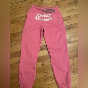 Pink sweatpants with white writing “little bit dramatic” with green string ties
